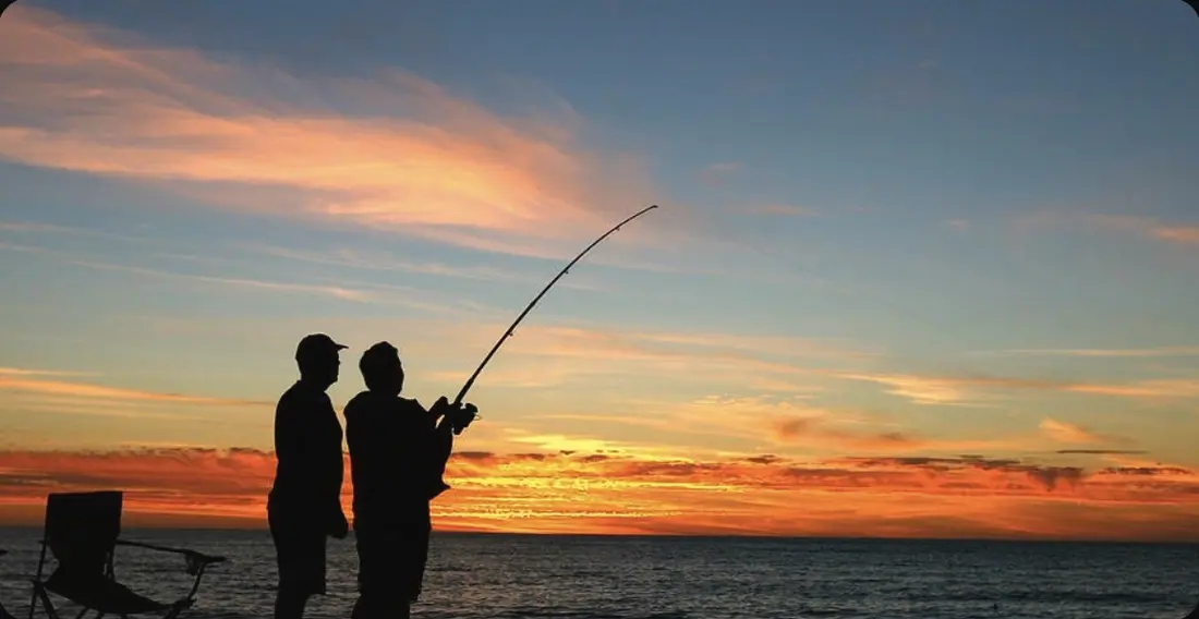Two friends fishing on the beach at sunset